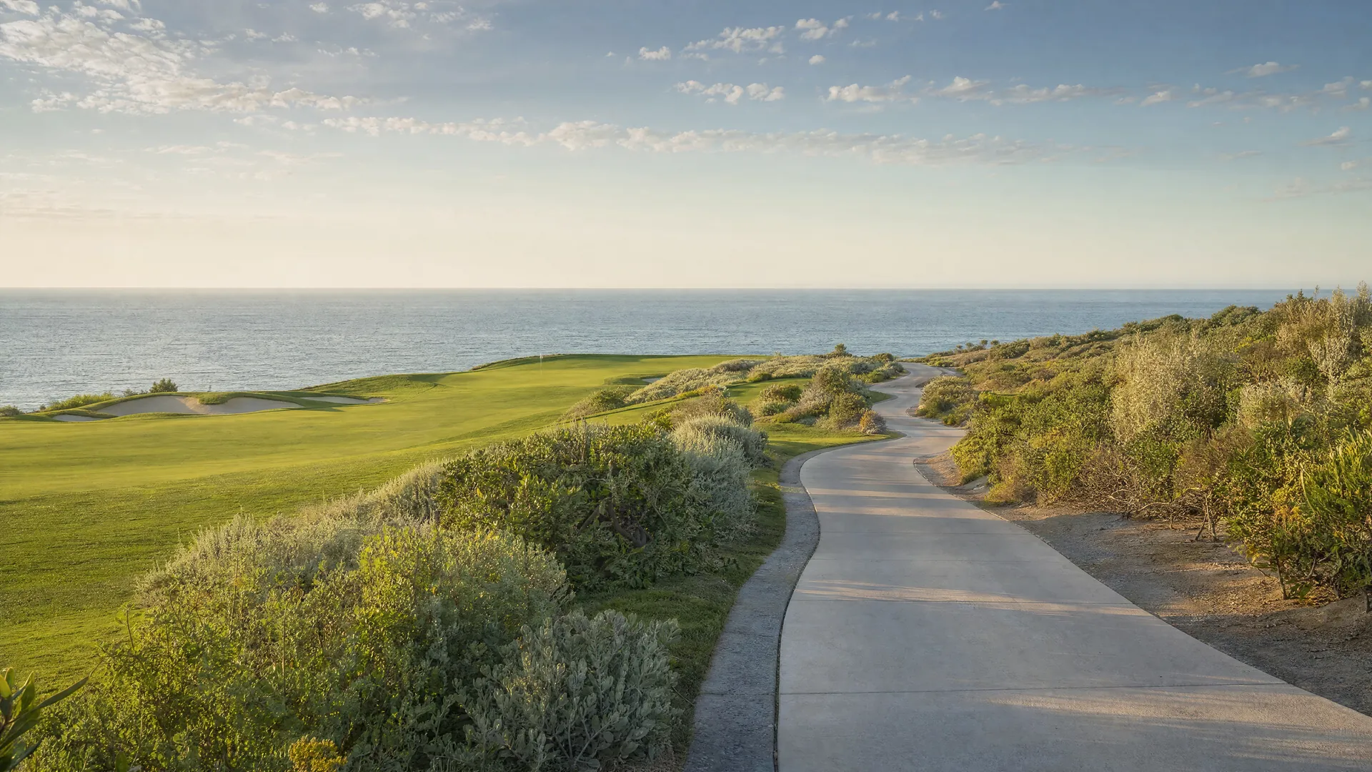 An empty concrete path winding through a golf fairway and desert scrub with the sea beyond