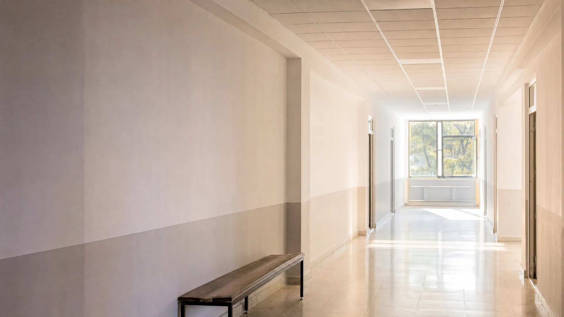 A bright empty corridor seen from afar with a bench and daylight from a window at the far end