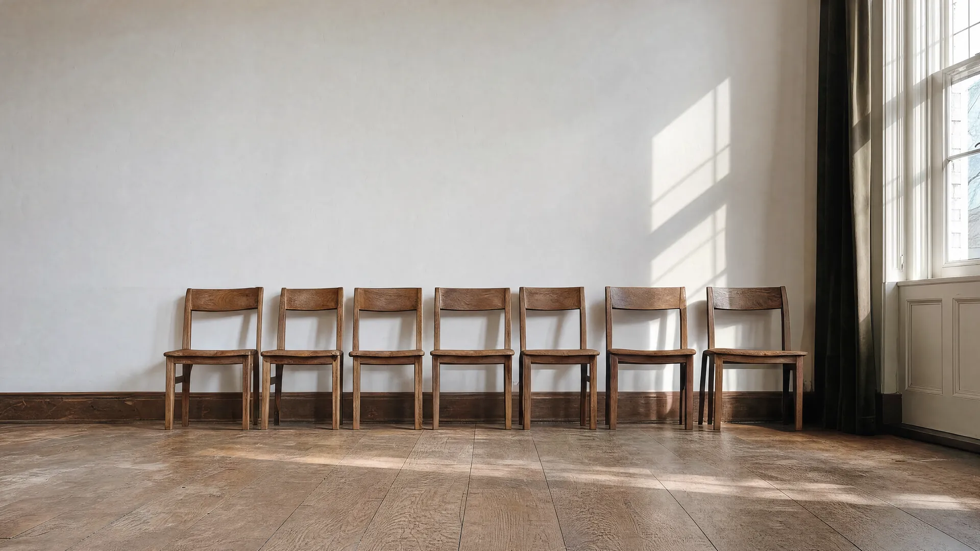 Una fila de sillas de madera vacías junto a un muro claro en una sala luminosa